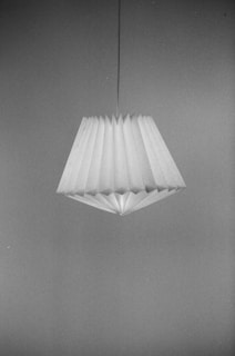 A pleated lampshade with a geometric design hangs from a cord against a plain background. The image is in black and white, highlighting the textures and folds of the shade.