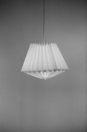 A pleated lampshade with a geometric design hangs from a cord against a plain background. The image is in black and white, highlighting the textures and folds of the shade.