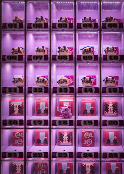 A grid of illuminated vending machine compartments contains a selection of colorful figurines and boxes. Each compartment is numbered from 01 to 25 and features lighting that emits a pinkish glow. The top row contains pug-looking figurines, while other rows display small red boxes with Chinese characters and some featuring a small dragon figurine.