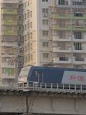A blue and white train is traveling on a raised track in front of a residential building. The building features multiple balconies with metal grilles and some windows have air conditioning units. The structure seems densely packed and the lighting suggests a calm, possibly late afternoon setting.