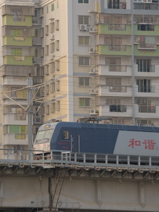 A blue and white train is traveling on a raised track in front of a residential building. The building features multiple balconies with metal grilles and some windows have air conditioning units. The structure seems densely packed and the lighting suggests a calm, possibly late afternoon setting.