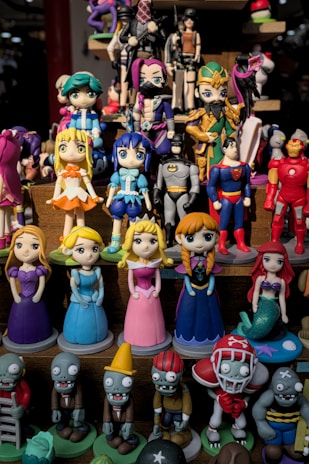A collection of colorful figurines displayed on shelves, featuring various characters in different costumes including princesses, superheroes, and zombies. The figurines are made in a playful, cartoonish style.