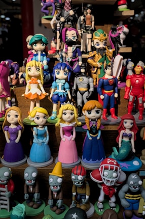 A collection of colorful figurines displayed on shelves, featuring various characters in different costumes including princesses, superheroes, and zombies. The figurines are made in a playful, cartoonish style.