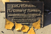 A metal plaque titled 'The Statue of Harmony' with an inscription in both Czech and English, surrounded by several yellow autumn leaves. The surface of the plaque has a weathered appearance, suggesting exposure to the elements.