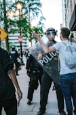 A tense street scene with a police officer holding a shield labeled 'POLICE' confronting a man wearing glasses and a backpack. The officer is wearing a helmet and visor, and there are other police officers in the background. The environment includes trees, buildings, and street signs.