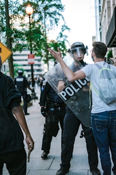 A tense street scene with a police officer holding a shield labeled 'POLICE' confronting a man wearing glasses and a backpack. The officer is wearing a helmet and visor, and there are other police officers in the background. The environment includes trees, buildings, and street signs.