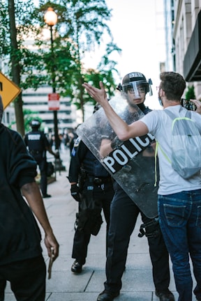 A tense street scene with a police officer holding a shield labeled 'POLICE' confronting a man wearing glasses and a backpack. The officer is wearing a helmet and visor, and there are other police officers in the background. The environment includes trees, buildings, and street signs.