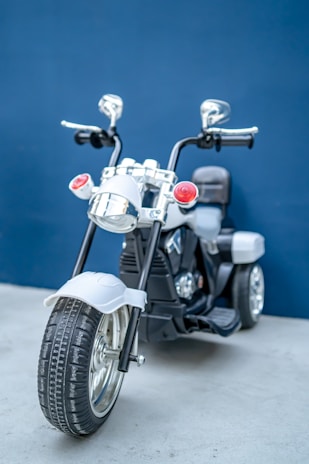 A small, sleek electric toy motorcycle with shiny chrome details and a black and white color scheme. The motorcycle has two rear-view mirrors, red lights, and a wide front tire, positioned in front of a blue background.