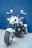 A small, sleek electric toy motorcycle with shiny chrome details and a black and white color scheme. The motorcycle has two rear-view mirrors, red lights, and a wide front tire, positioned in front of a blue background.