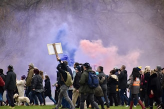 A group of people are gathered in an outdoor setting, surrounded by colorful smoke in shades of purple and orange. Some individuals are holding signs, and a small dog is visible among the crowd. The atmosphere is dynamic and possibly related to a demonstration or public gathering.