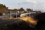 A high-speed white train travels on railway tracks alongside a road with moving vehicles, including cars and trucks. The scene is set in a rural area with sparse trees and vegetation. Two large wind turbines stand in the background, towering over the landscape. The sun casts long shadows, suggesting it is either early morning or late afternoon.