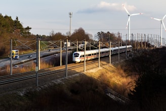 A high-speed white train travels on railway tracks alongside a road with moving vehicles, including cars and trucks. The scene is set in a rural area with sparse trees and vegetation. Two large wind turbines stand in the background, towering over the landscape. The sun casts long shadows, suggesting it is either early morning or late afternoon.