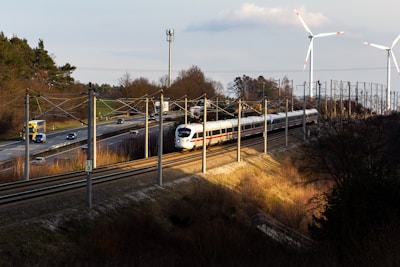 A high-speed white train travels on railway tracks alongside a road with moving vehicles, including cars and trucks. The scene is set in a rural area with sparse trees and vegetation. Two large wind turbines stand in the background, towering over the landscape. The sun casts long shadows, suggesting it is either early morning or late afternoon.