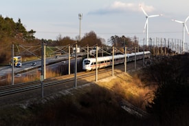 A high-speed white train travels on railway tracks alongside a road with moving vehicles, including cars and trucks. The scene is set in a rural area with sparse trees and vegetation. Two large wind turbines stand in the background, towering over the landscape. The sun casts long shadows, suggesting it is either early morning or late afternoon.