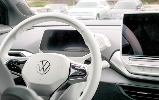 The image captures the interior of a modern car, highlighting a steering wheel with a Volkswagen logo, a digital display screen, and a sleek dashboard design. The surrounding environment is visible through the windshield, showing parked cars outside.