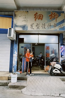 A woman in a red jacket stands outside a hair salon, holding a phone while a motorbike is parked nearby. The salon has a large sign with Chinese characters and an artistic image of a face on it. The entrance is open, revealing a person getting a haircut inside.