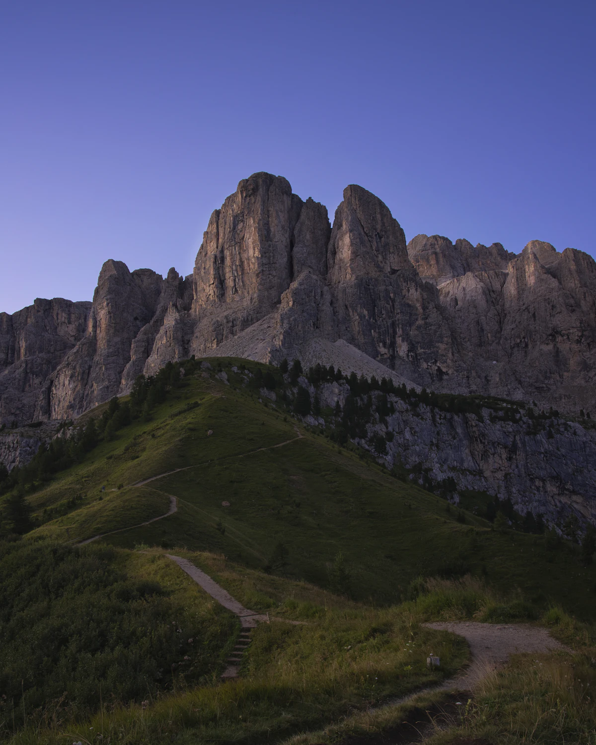 A gravel track climbing a grassy shoulder below a tall rock wall near Passo Gardena, at dawn