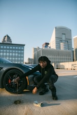 A person in dark clothing is crouched next to a car wheel on a rooftop parking lot. The car appears sporty, and there's a stack of cash on the ground. In the background, there is a cityscape with tall modern buildings.
