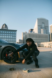 A person in dark clothing is crouched next to a car wheel on a rooftop parking lot. The car appears sporty, and there's a stack of cash on the ground. In the background, there is a cityscape with tall modern buildings.
