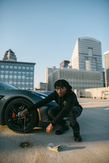 A person in dark clothing is crouched next to a car wheel on a rooftop parking lot. The car appears sporty, and there's a stack of cash on the ground. In the background, there is a cityscape with tall modern buildings.