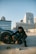 A person in dark clothing is crouched next to a car wheel on a rooftop parking lot. The car appears sporty, and there's a stack of cash on the ground. In the background, there is a cityscape with tall modern buildings.