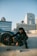 A person in dark clothing is crouched next to a car wheel on a rooftop parking lot. The car appears sporty, and there's a stack of cash on the ground. In the background, there is a cityscape with tall modern buildings.