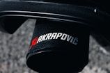 A close-up image of a black cylindrical exhaust tip featuring the branding logo of Akrapovič in red and silver letters. The background consists of a blurred, textured surface, possibly concrete or asphalt.