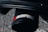 A close-up image of a black cylindrical exhaust tip featuring the branding logo of Akrapovič in red and silver letters. The background consists of a blurred, textured surface, possibly concrete or asphalt.