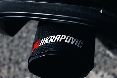 A close-up image of a black cylindrical exhaust tip featuring the branding logo of Akrapovič in red and silver letters. The background consists of a blurred, textured surface, possibly concrete or asphalt.