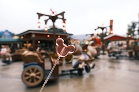 The image focuses on a pink lollipop shaped like a famous mouse head, held up against a blurry background of an amusement park or theme park. The park features a red building, festive lights, wooden carts, and people walking around, creating a lively atmosphere.