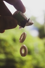 A hand holds a partially unwrapped roll of mint candies, with two individual candies falling through the air. The background is a blurred, lush green, suggesting an outdoor setting.