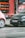 Two cars are parked on a street beside a small market stall filled with crates of oranges and other produce. The background features a red wall with advertisements, and the scene appears to be part of an urban setting.