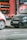 Two cars are parked on a street beside a small market stall filled with crates of oranges and other produce. The background features a red wall with advertisements, and the scene appears to be part of an urban setting.
