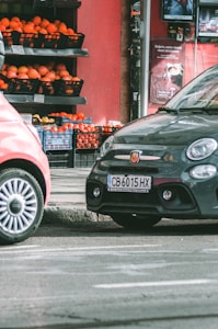 Two cars are parked on a street beside a small market stall filled with crates of oranges and other produce. The background features a red wall with advertisements, and the scene appears to be part of an urban setting.