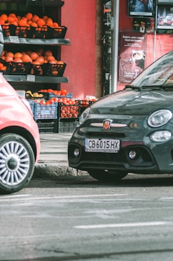 Two cars are parked on a street beside a small market stall filled with crates of oranges and other produce. The background features a red wall with advertisements, and the scene appears to be part of an urban setting.