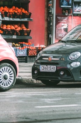 Two cars are parked on a street beside a small market stall filled with crates of oranges and other produce. The background features a red wall with advertisements, and the scene appears to be part of an urban setting.
