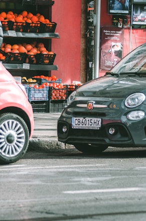 Two cars are parked on a street beside a small market stall filled with crates of oranges and other produce. The background features a red wall with advertisements, and the scene appears to be part of an urban setting.
