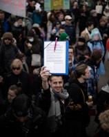 A man is holding up a clipboard with a document attached, surrounded by a crowd of people. Many individuals are dressed warmly, some wearing hats and scarves, and a few are holding signs. The gathering appears to be a public demonstration or protest.