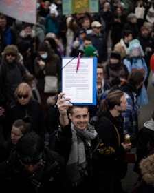 A man is holding up a clipboard with a document attached, surrounded by a crowd of people. Many individuals are dressed warmly, some wearing hats and scarves, and a few are holding signs. The gathering appears to be a public demonstration or protest.