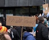 A crowd of people gathered, holding signs. One prominent cardboard sign reads 'Ich will Party!' in a handwritten style. The crowd is dressed in winter clothing, indicating a colder climate. The background is urban, with a dark building façade visible.