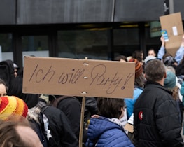 A crowd of people gathered, holding signs. One prominent cardboard sign reads 'Ich will Party!' in a handwritten style. The crowd is dressed in winter clothing, indicating a colder climate. The background is urban, with a dark building façade visible.