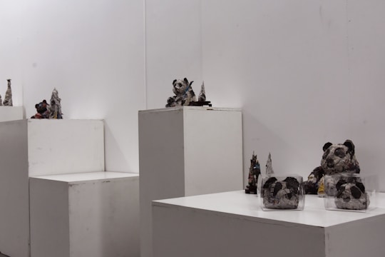 Several small sculptures are displayed on white pedestals in a minimalist gallery space. The sculptures appear to be crafted from recycled materials, resembling abstract or animal-like forms, including figures that look like pandas. The background is plain and white, drawing attention to the textures and details of the sculptures.