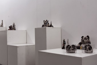 Several small sculptures are displayed on white pedestals in a minimalist gallery space. The sculptures appear to be crafted from recycled materials, resembling abstract or animal-like forms, including figures that look like pandas. The background is plain and white, drawing attention to the textures and details of the sculptures.