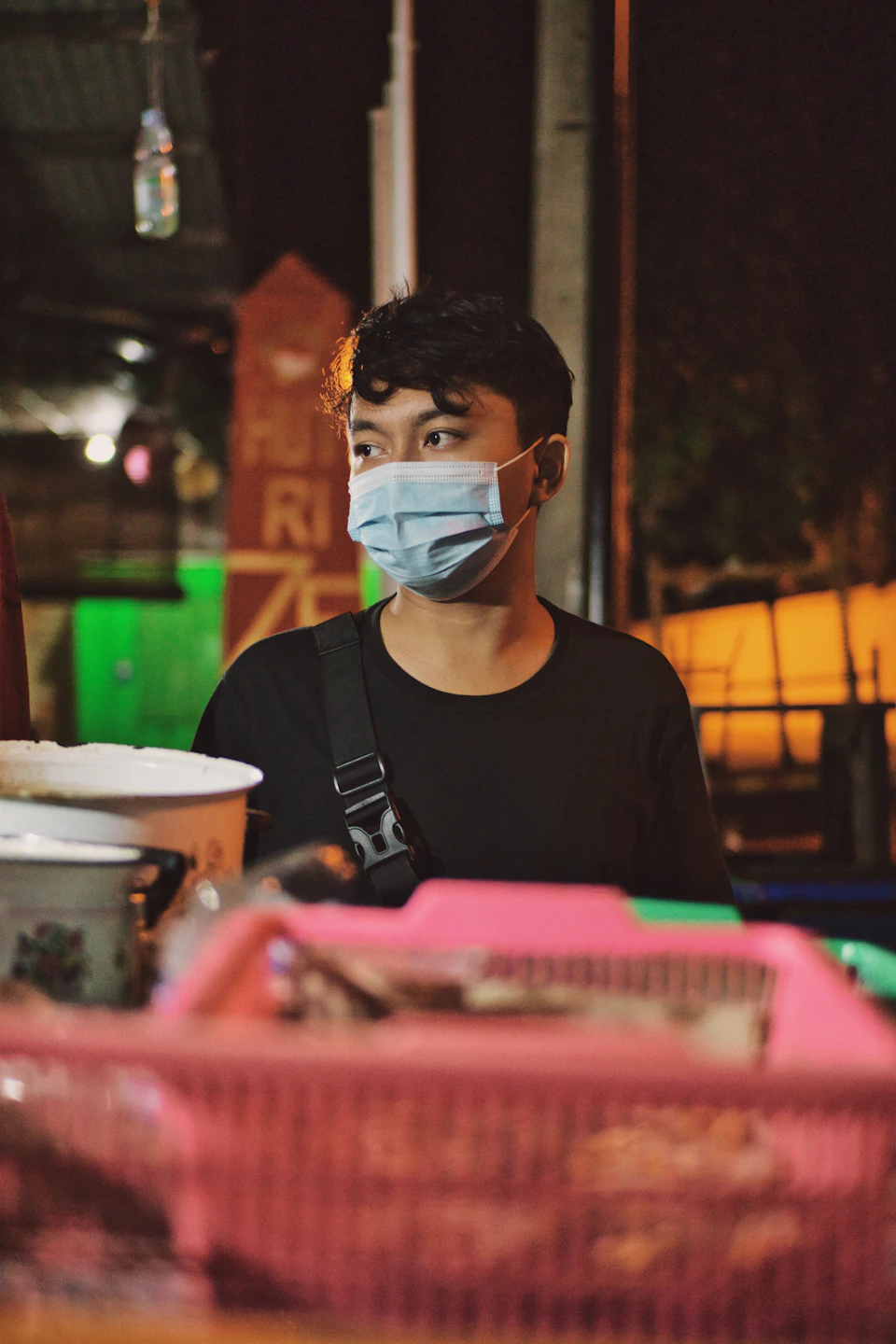 Young man wearing face mask stands beside table with scattered items, gazing outward