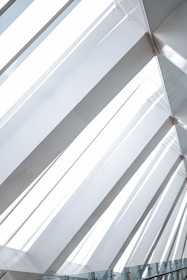 Large geometric glass panels form an angled skylight, allowing natural light to flood the space. The structure features clean lines and a modern architectural design, emphasizing transparency and openness. Reflections and shadows create subtle patterns on the glass surface.