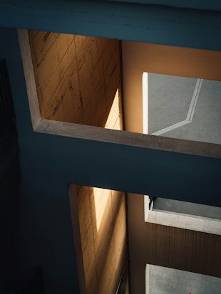 A modern architectural structure with a geometric design featuring angular beams and shadows. The interplay of light and shadow creates a dramatic effect on the textured walls and surfaces.
