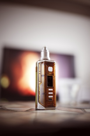 A sleek, metallic vape device stands on a table with a warm, blurred background. The device has a transparent section revealing some internal components and a digital display. Soft lighting highlights the contours and shiny surfaces.