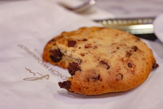 A chocolate chip cookie with a bite taken out of it is placed on a white paper with some text printed on it. In the background, there is a blurred view of a knife and a white plate.