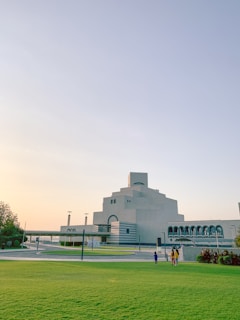 A modern architectural structure with clean lines and a geometric design dominates the background. The building is surrounded by a manicured green lawn with a few people walking towards it. The sky is clear with warm tones indicating either sunrise or sunset.