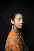 A young person with a braided hairstyle is wearing an ornate outfit that includes a grey and orange garment and intricate jewelry, including a dangle earring with a circular design and beads around the neck. The background is dark with a subtle geometric pattern.